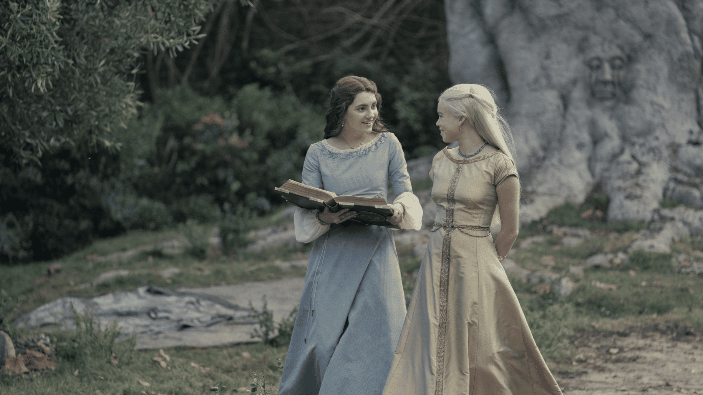 Two young women in medieval aristocratic garb chat over a book while standing in front of a tree with a face carved in it